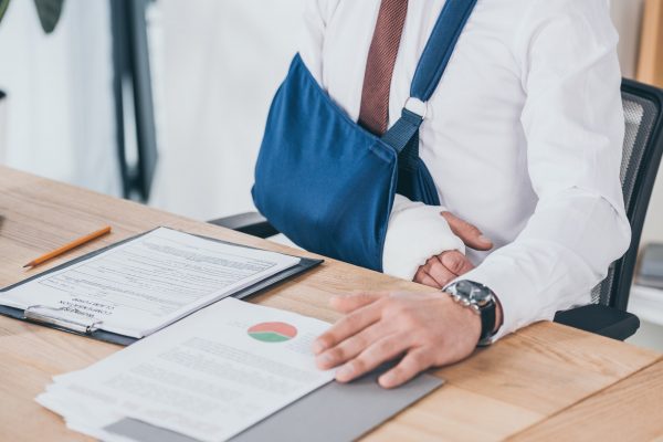 cropped view of worker with broken arm in bandage sitting at table in office, compensation concept